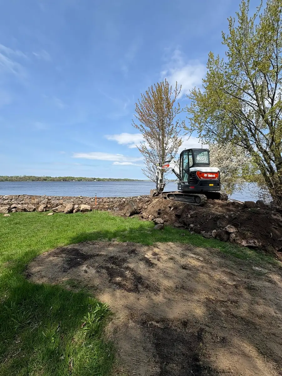 Excavatrice compacte de l'Entreprise SME au travail au bord d'un plan d'eau, terre fraîchement remuée devant l'enrochement