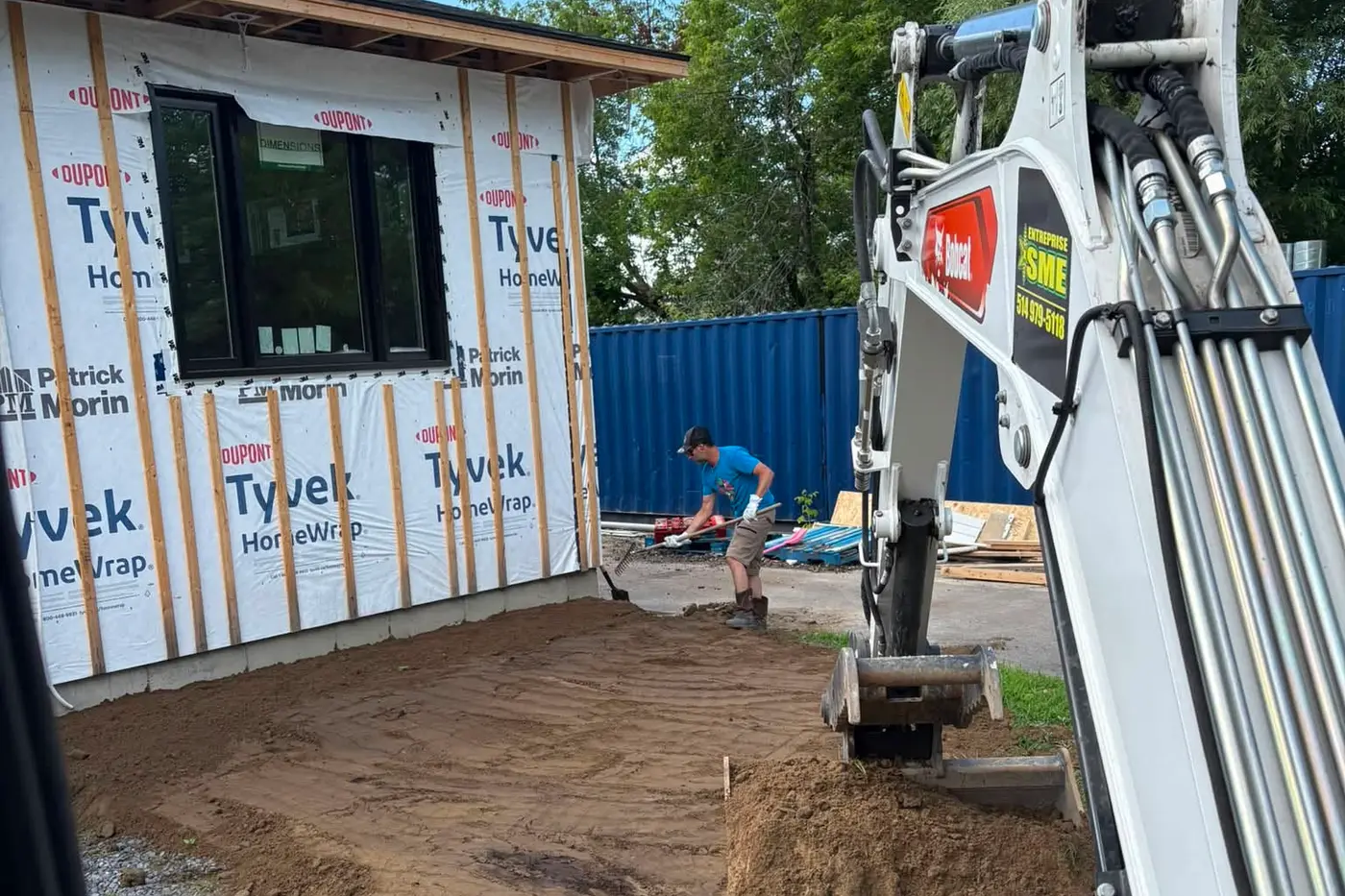 Pelle compacte de l'Entreprise SME au travail contre une maison en construction, godet chargé de terre